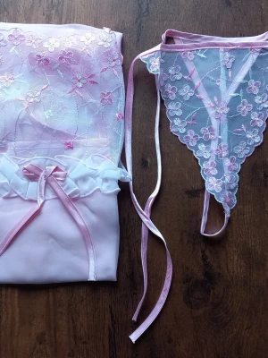 Rosa spetsbody med trosa från Enigma storlek M - Sött set från Enigma med en ljusrosa body i mesh och spets, dekorerad med blommiga broderier och vita volanger. Matchande stringtrosa med samma spetsdetaljer och tunna band. Perfekt för dig som gillar romantiska och feminina underkläder. Storlek M 