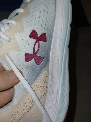 Under Armour vita sneakers - Snygga vita sneakers från Under Armour med beige detaljer och röd logga på sidan. Ovandelen är i mesh för bra andningsförmåga och sulan är vit med Charged Cushioning för extra komfort. Perfekta för dig som gillar sportig stil och vill ha bekväma skor till vardags.