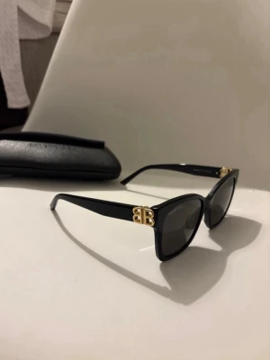 Svarta Balenciaga solglasögon med guldlogo - Snygga svarta solglasögon från Balenciaga med klassisk cat eye-form och guldig BB-logo på sidorna. Glasögonen har mörka linser och kommer med originalfodral. Perfekt accessoar för att lyfta din stil.