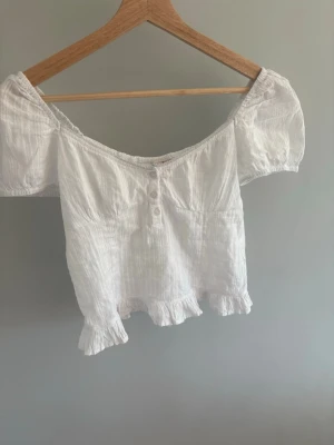 Vit offshoulder blus med volang - Söt vit blus i bomull med offshoulder-modell och korta puffärmar. Toppen har knappar framtill, smockad rygg och volangkant nertill. Perfekt för en somrig och avslappnad look. Använd en gång!