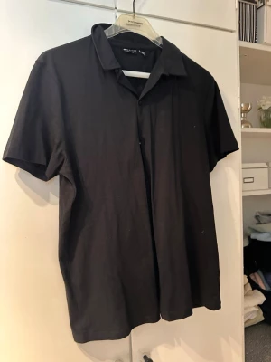 Svart kortärmad skjorta - En stilren svart skjorta med korta ärmar och klassisk krage. Skjortan är i mjukt bomullsmaterial och har en avslappnad passform. Perfekt för en clean och enkel look.