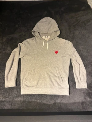 Grå hoodie från Comme des Garçons Play - Snygg grå hoodie med huva och dragsnören från Comme des Garçons Play. Har det ikoniska röda hjärtat broderat på bröstet och en stor magficka. Perfekt för en chill och stilren look. Är mindre i storleken passar mer åt storlek S