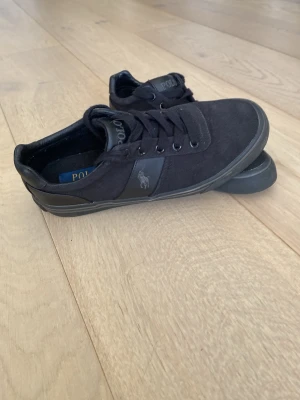 Svarta Polo Ralph Lauren sneakers - Snygga svarta sneakers från Polo Ralph Lauren med klassisk låg profil. Skorna har textilovandel, svarta snören och detaljer i svart läder vid häl och sidor. Polo-logga på sidan och bak på sulan. Perfekta för en clean och stilren look.