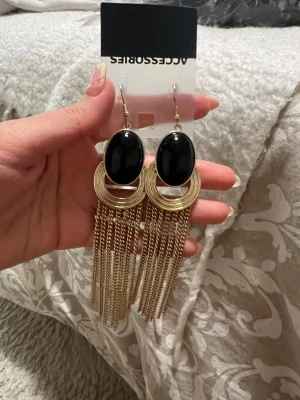 Svart-guldiga statementörhängen Accessorize - Snygga statementörhängen från Accessorize med stora svarta ovala stenar, guldfärgade ringar och långa kedjor som hänger ner. Perfekta för att lyfta vilken outfit som helst med sin coola design och mix av svart och guld. Krokar för enkel användning.