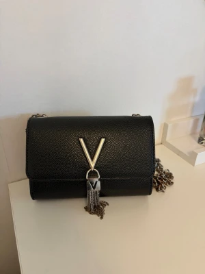 Svart handväska från Valentino - Snygg svart handväska från Valentino med kedjerem och ett stort V i silver framtill. Väskan har en stilren, rektangulär form och är gjord i ett strukturerat lädermaterial. Silverdetaljer och dekorativa fransar framtill ger en lyxig känsla.