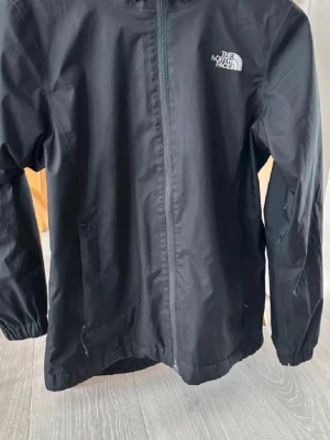  The North Face Windbreaker - Svart vindjacka från The North Face med huva och dragkedja framtill. Jackan har två sidofickor med dragkedja och elastiska muddar vid ärmsluten. Lite schysstare jacka och enkel att matcha med andra plagg.
