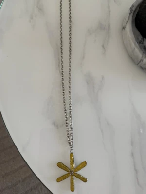 Silverfärgat halsband med gul blomma - Unikt halsband med en tjock silverfärgad kedja och ett stort hängsmycke i form av en blomma med sex gula kronblad. Blomman har en retrovibe och är tillverkad i metall med emaljerade detaljer. Perfekt statement piece för dig som gillar att sticka ut.