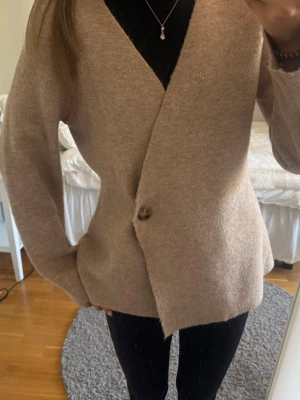 Beige kofta från Neo Noir - Mysig beige kofta från Neo Noir med v-ringning och en knapp framtill. Koftan har en avslappnad passform och är tillverkad i ett mjukt, stickat material som känns skönt mot huden. Perfekt att slänga på sig över en topp eller t-shirt. Storlek står ej, men jag är ca 1,69 cm