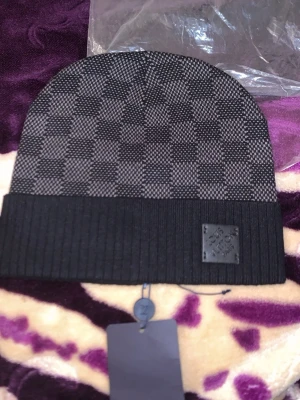 Louis Vuitton svart och grå mössa - Snygg svart och grå mössa från Louis Vuitton med klassiskt rutmönster och ribbad uppvikt kant. Märkeslogga i svart läder framtill. Perfekt accessoar för att lyfta din streetstyle. Materialet ser ut att vara stickat och känns lyxigt.