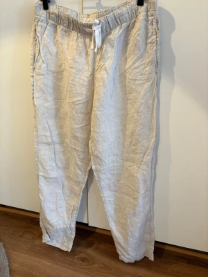 Beiga linnebyxor med dragsko - Snygga och luftiga beiga linnebyxor med resår och vit dragsko i midjan. Byxorna har raka ben och sidofickor, perfekta för en avslappnad och trendig stil. 