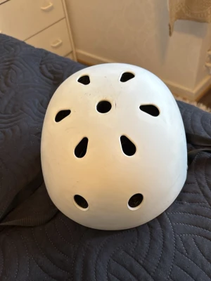 Vit hjälm - Säljer en vit hjälm med flera ventilationshål och justerbart spänne. Hjälmen har svart innerfoder och remmar för säker passform. Passar till cykling, skateboard eller andra aktiviteter där huvudskydd behövs.