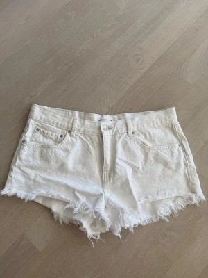 Vita jeansshorts med fransig kant - Säljer ett par vita jeansshorts från Gina Tricot med fransig kant nertill. Klassisk femficksmodell med knapp och dragkedja framtill. Perfekta för sommaren och har en avslappnad, trendig look.