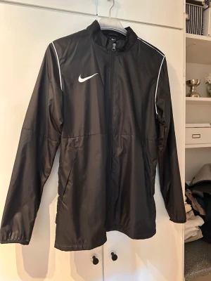 Svart Nike vindjacka med vita detaljer - Snygg svart vindjacka från Nike med vita detaljer och dragkedja framtill. Jackan har hög krage, långa ärmar och tunnt, lätt material som passar perfekt för blåsiga dagar. Ikonisk Nike-logga på bröstet och vita piping längs axlarna ger en sportig look.