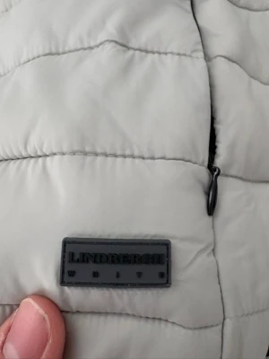 Ljusgrå dunväst från Lindbergh - Snygg ljusgrå dunväst från Lindbergh med quiltad design och svart dragkedja. Västen har en hög krage och svarta detaljer vid ärmöppning och nederkant. Perfekt för lager-på-lager och enkel att matcha med olika outfits.