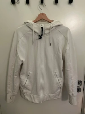 Vit C.P. Company kofta - Vit hoodie med hel dragkedja och två fickor med dragkedja framtill. Klassisk C.P. Company-design med svarta goggles i huvan och justerbara snören. Perfekt för dig som gillar streetwear och unika detaljer. Finns ett litet hål på ena armen. Köpt i NK men tappat bort kvittot 