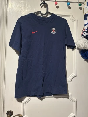 PSG t-shirt med nummer 10 - Mörkblå PSG t-shirt från Nike med klubbmärke och röd swoosh på bröstet. På ryggen finns siffran 10 tryckt i vitt och en liten fransk flagga. Perfekt för fotbollsfans som vill visa sitt stöd för Paris Saint-Germain.