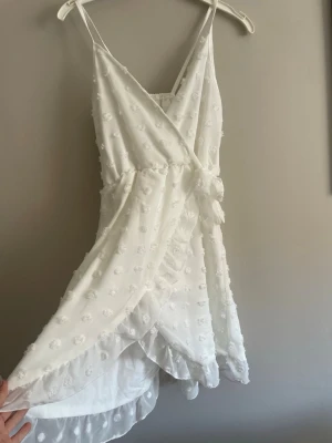 Vit omlottklänning med volang - Superfin vit omlottklänning med smala axelband och volangkant. Klänningen har knyt i midjan och är dekorerad med små broderade blommor. Perfekt för sommarens alla tillfällen och ger en romantisk vibe. Stl 11-12 år men jag som har xs passar den bra, jag är 168 cm