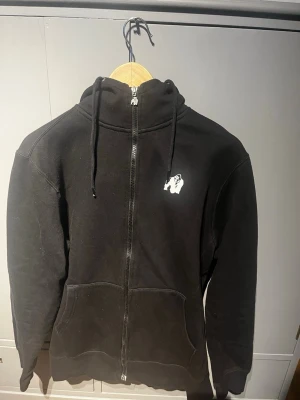 Svart zip hoodie från Gorilla Wear - Säljer en svart hoodie med dragkedja från Gorilla Wear. Hoodien har huva med dragsko, fickor framtill och en liten vit logga på bröstet. Tillverkad i mjukt material som känns skönt mot huden. Storlek M