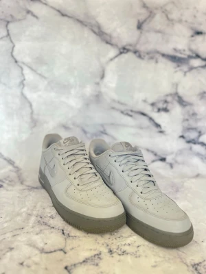 Nike Air Force 1  sneakers - Klassiska Nike Air Force 1 sneakers i helvitt läder med matchande vita skosnören och grå sula. Ikonisk swoosh-logga på sidorna och perforerade detaljer på tån för extra ventilation. Perfekta för en clean och stilren look.                                                                                  Priset kan diskuteras!