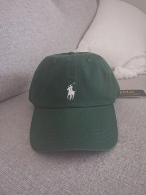 Mörkgrön keps från Polo Ralph Lauren - Snygg mörkgrön keps från Polo Ralph Lauren med klassisk broderad logga framtill och 'POLO' broderat baktill. Justerbar rem baktill för perfekt passform. Tillverkad i bomullstyg som känns både stilrent och bekvämt.