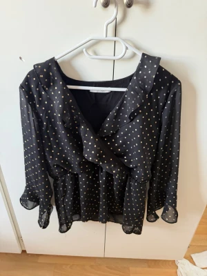 Svart blus med guldprickar från Vila - Snygg svart blus från Vila med guldiga prickar och volangdetaljer framtill. Blusen har långärmad design och är gjord i ett tunt, transparent material som ger en luftig känsla. Perfekt att styla med jeans eller kjol för en trendig look.