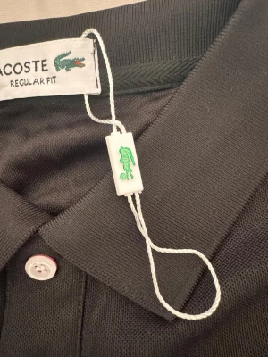 Svart pikétröja från Lacoste M - Klassisk svart pikétröja från Lacoste med regular fit. Tröjan har korta ärmar, krage och knappar framtill. Tillverkad i bomull för en bekväm känsla och har den ikoniska gröna krokodilloggan på bröstet.