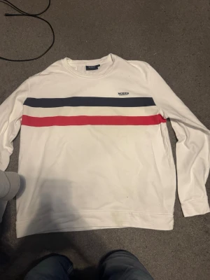 Vit sweatshirt från Morris Company - Vit sweatshirt med rund hals från Morris Company. Tröjan har två horisontella ränder över bröstet, en marinblå och en röd, samt en liten broderad logga på vänster sida. Tillverkad i mjukt bomullsmaterial och har långa ärmar.