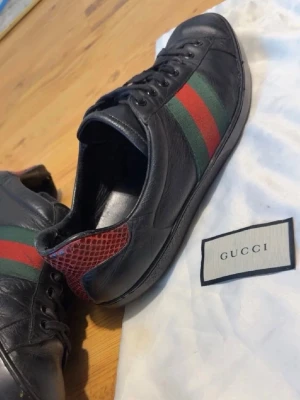 Svarta Gucci sneakers med röd/grön rand - Snygga svarta sneakers från Gucci i läder med klassiska gröna och röda ränder på sidan. Hälpartiet har en röd detalj med ormmönster som ger extra edge. Skorna har lågt skaft och svart snörning. Perfekta för dig som vill ha en lyxig och trendig look.