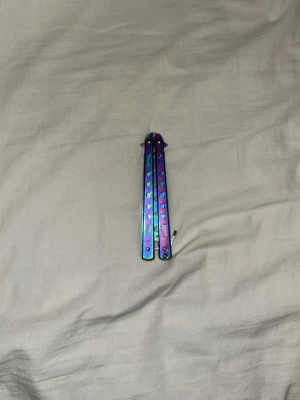 Regnbågsfärgad butterfly-träningskniv - Cool butterfly-träningskniv i metall med regnbågsfärgad finish. Handtagen har hål för bättre grepp och kniven är designad för tricks och övning, inte skarp. Perfekt för dig som vill lära dig flip tricks eller bara gillar snygga accessoarer.