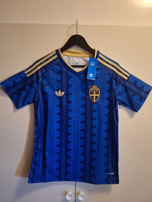 Sverige VM 2026 fotbollströja  - Blå fotbollströja från Adidas med Sveriges landslagsemblem och guldiga detaljer. Tröjan har ett unikt mönster, korta ärmar och klassiska Adidas-ränder på axlarna. Perfekt för dig som vill sticka ut på läktaren eller träningen.
