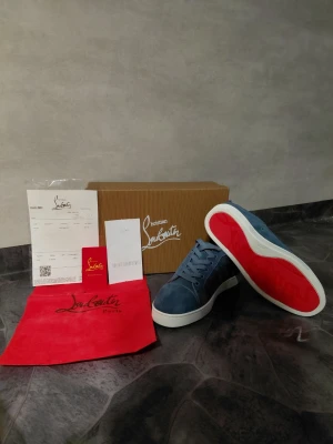 Blå Christian Louboutin sneakers - Snygga blå sneakers från Christian Louboutin med klassisk röd sula och vita kanter. Skorna har lågt skaft, snörning och är tillverkade i mocka. Perfekta för dig som vill sticka ut med exklusiva detaljer och ikonisk design.