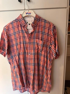 Rutig kortärmad skjorta i rött och blått - Kortärmad skjorta med rött, blått och vitt rutmönster. Klassisk krage med knappar och bröstficka. Perfekt för en avslappnad och stilren look. Skjortan är i bomull och har en normal passform.