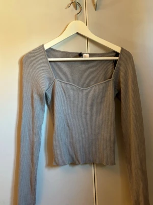 Ljusgrå ribbad långärmad topp - Supersnygg ljusgrå ribbad topp med lång ärm och v-ringad skärning framtill. Figurnära passform och stretchigt material som sitter bekvämt. Perfekt att styla med jeans eller kjol för en trendig look.