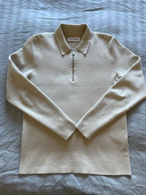 Beige halvzip-tröja från Samsøe Samsøe - Stilren beige tröja från Samsøe Samsøe med halv dragkedja och klassisk krage. Tröjan har lång ärm och är tillverkad i ett mjukt, strukturerat material som ger en modern look. Perfekt för en clean och avslappnad stil.