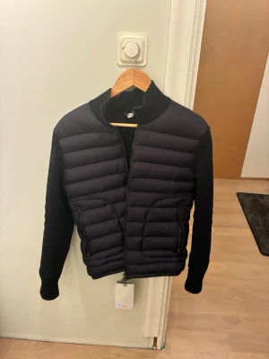  Monclercardigan - Svart cardiganjacka från Moncler med quiltad front och stickade ärmar. Jackan har dragkedja framtill, två sidofickor med dragkedja och hög krage. Moncler-logga på ärmen för en exklusiv vibe. Perfekt för dig som gillar stilren och modern design.