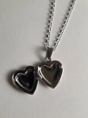 Silverfärgat hjärthalsband med medaljong - Säljer ett silverfärgat halsband med hjärtformad medaljong som går att öppna. Kedjan är tunn i rostfritt stål och medaljongen har fina detaljer runt kanten. Perfekt för att lägga i ett litet foto eller minne. Materialet ser ut som metall och designen är stilren och klassisk.