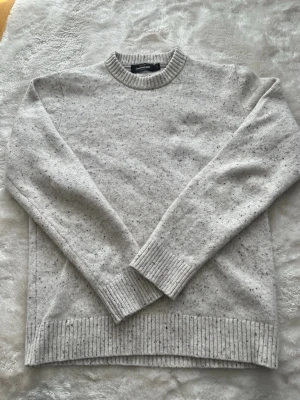 Ljusgrå stickad tröja från Dressmann - Säljer en ljusgrå stickad tröja från Dressmann med diskreta mörkare prickar i materialet. Tröjan har rund halsringning, ribbade muddar vid ärmslut och nederkant samt en klassisk rak passform. Perfekt för lager på lager under kyliga dagar.