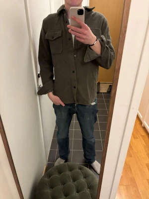 Olivgrön overshirt från Dobber - Säljer en olivgrön overshirt från Dobber med två stora bröstfickor och knappar framtill. Overshirten har klassisk krage, långa ärmar och är gjord i ett mjukt, lite tjockare tyg som passar perfekt till lager på lager-stil. EN KNAPP ÄR AV