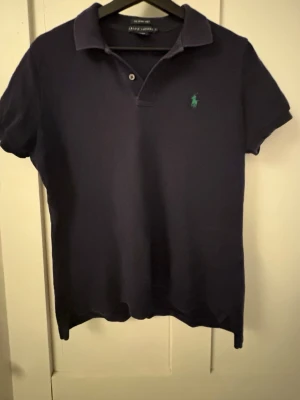Marinblå pikétopp från Ralph Lauren - Snygg marinblå pikétopp från Ralph Lauren med klassisk krage, korta ärmar och grön broderad logga på bröstet. Perfekt tröja nu mår vår/sommar. Står att tröjan är i L men de är väldigt liten och passar egentligen S