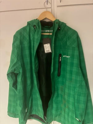 Grön vindjacka från Everest - Grön vindjacka från Everest i rutigt mönster. Jackan har huva med dragsko, bröstficka med dragkedja och meshfoder på insidan. Perfekt för blåsiga dagar och outdoor-aktiviteter. Stängs med dragkedja framtill och har justerbara ärmslut.