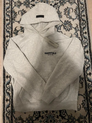 Grå Essentials Fear of God hoodie - Snygg ljusgrå hoodie från Essentials Fear of God med svart logga på bröstet och en diskret patch på huvan. Klassisk passform med känguruficka och ribbade muddar. Tillverkad i mjuk bomullsblandning för maximal komfort.