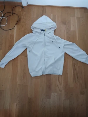 Vit hoodie från Polo Ralph Lauren - Säljer en stilren vit hoodie från Polo Ralph Lauren med dragkedja framtill och klassisk huva med snören. Tröjan har två fickor på magen och den ikoniska lilla loggan broderad på bröstet. Perfekt för en avslappnad och trendig look.