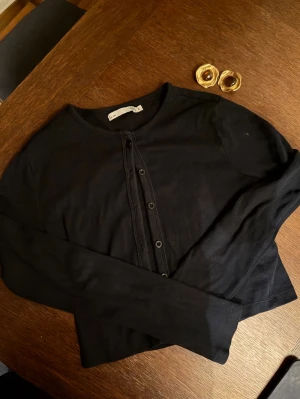 Svart ribbad kofta från Lager 157  - Säljer en svart ribbad kofta från Lager 157 med rund halsringning och knappar framtill. Koftan har lång ärm och en croppad passform som passar perfekt till jeans eller kjol. Enkel och stilren design som är lätt att matcha.