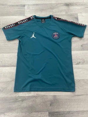 PSG x Jordan  - träningströja från PSG x Jordan med vita loggor på bröstet och Paris Saint-Germain-märke. Svarta axelband med vit text 'Jordan' längs ärmarna. 