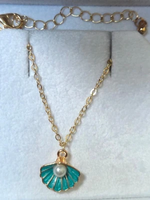 Halsband med blå snäcka och pärla - Snyggt halsband med tunn guldfärgad kedja och ett hängsmycke i form av en blå snäcka med en vit pärla i mitten. Perfekt accessoar för dig som gillar havsinspirerade detaljer. Kedjan är enkel och smidig, och snäckan har fina guldfärgade kanter.
