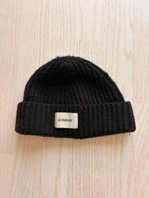 Svart ribbstickad mössa från Dondup - Säljer en svart ribbstickad mössa från Dondup med uppvikt kant och en beige logotyp-patch framtill. Mössan är gjord i ett mjukt, tjockt material som håller värmen och har en klassisk, enkel form som passar till det mesta.