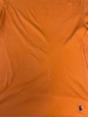 Orange t-shirt från Polo Ralph Lauren - Snygg orange t-shirt från Polo Ralph Lauren med klassisk passform och blå broderad logga längst ner. Tillverkad i 100% bomull för en skön känsla. Perfekt till jeans eller shorts för en avslappnad look.