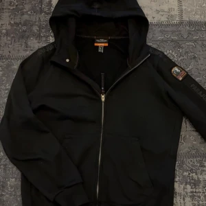 Svart hoodie från Parajumpers - Svart hoodie från Parajumpers med dragkedja framtill och stor huva. Snygg patch med logga på ena ärmen och diskret Parajumpers-tryck längs axeln. Mjuk bomullskänsla och två fickor framtill. Perfekt för en chill och stilren look.