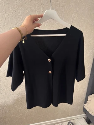 Snygg asymmetrisk topp från Shein – Storlek M - Säljer denna populära svarta topp i västmodell med en unik design. Den har en asymmetrisk knäppning framtill och dekorativa guldknappar som ger en lyxig look. Materialet är stretchigt och följsamt, vilket gör den väldigt bekväm att bära. • Märke: Shein • Storlek: M (38) • Skick: Mycket fint skick, endast använd två gånger. • Passform: Figurnära med stretch. Sitter perfekt till både högmidjade jeans eller kostymbyxor. Detaljer: • Snygga guldknappar som sticker ut. • Asymmetrisk skärning nertill 