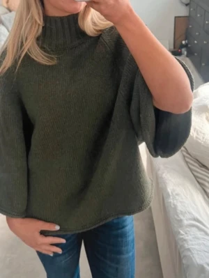 Olivgrön stickad poncho med polokrage - Säljer en olivgrön stickad poncho med hög polokrage och vida trekvartsärmar. Ponchon har en avslappnad passform och är perfekt för lager på lager. Materialet känns mjukt och bekvämt, troligen akryl eller ullblandning. Enkel och stilren design som passar till jeans.